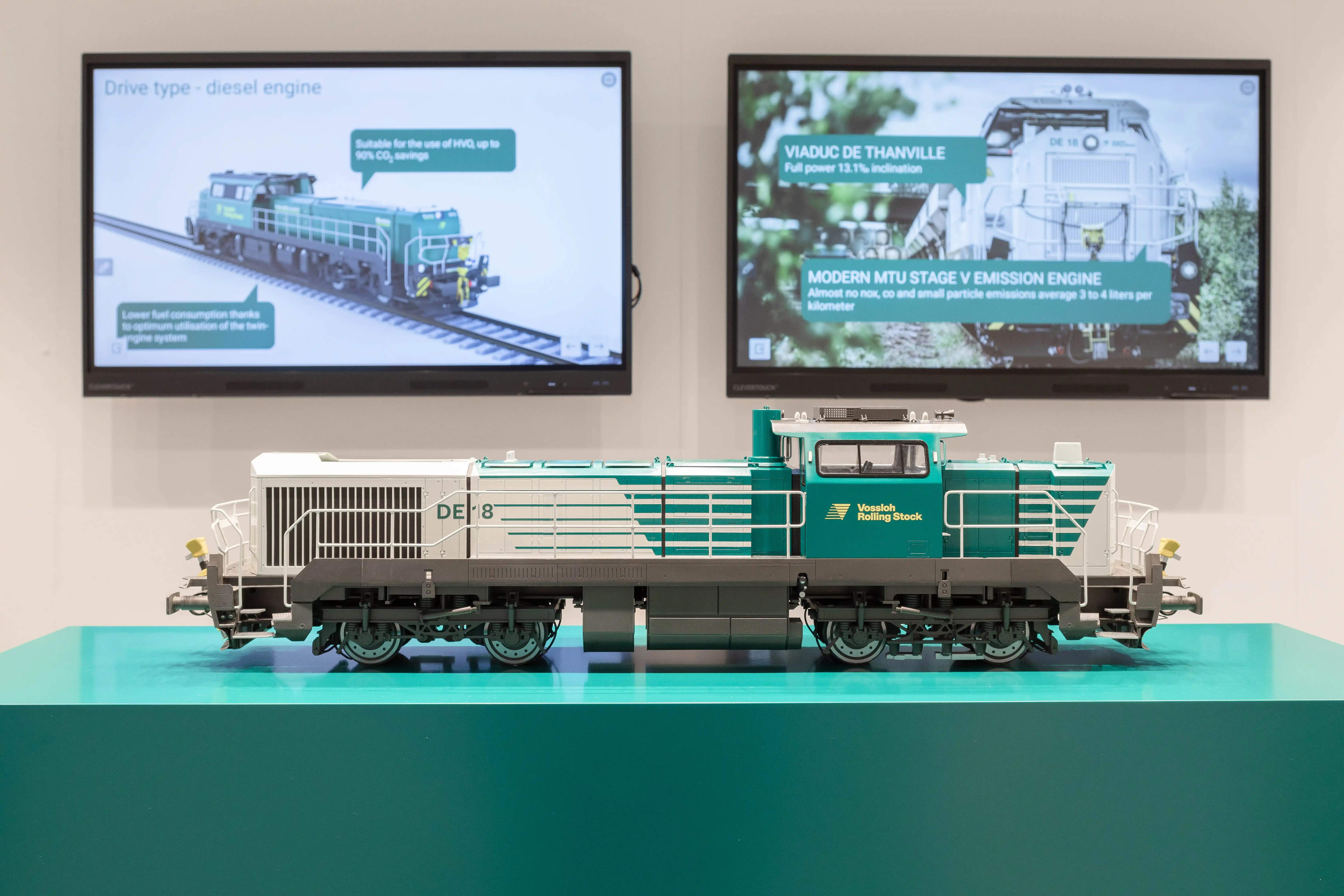 Detailed model of a green-white Vossloh diesel locomotive on a green plinth in front of two info screens.