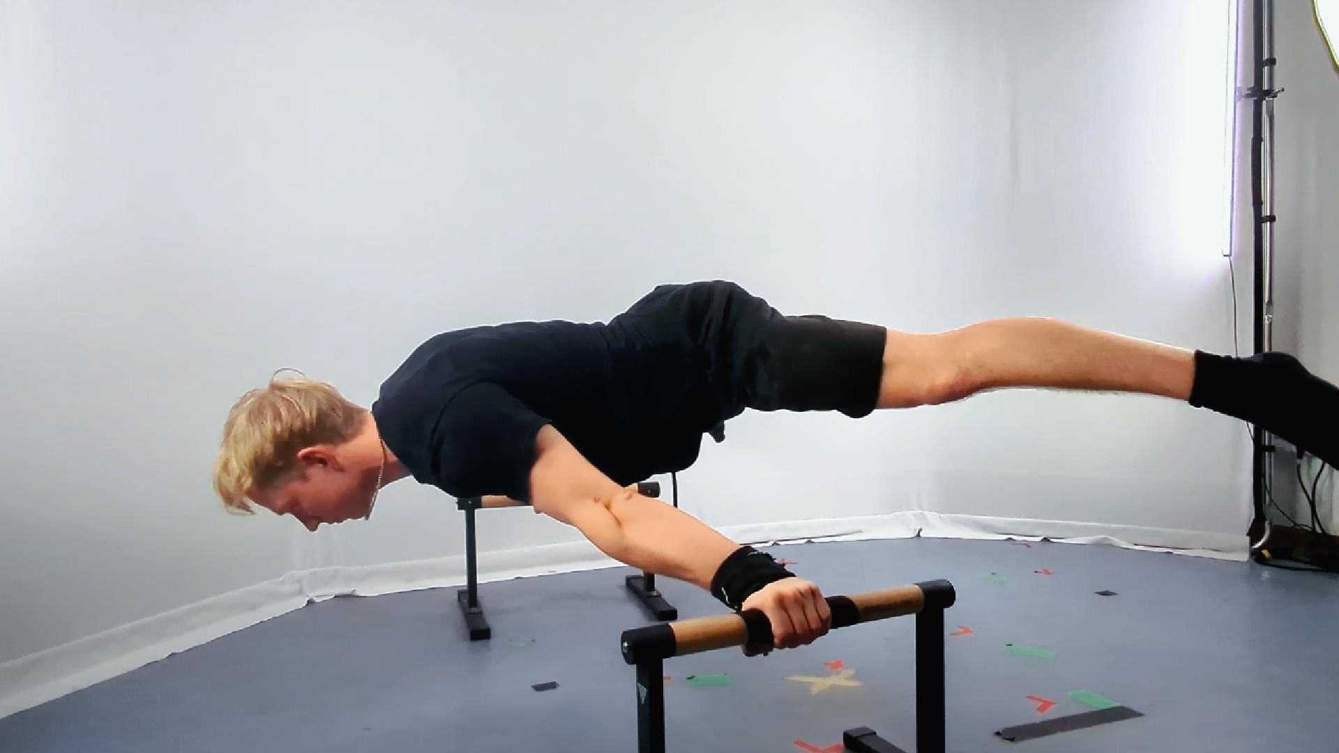 A person performs a Calisthenics Straddle Maltese hold, balancing horizontally on parallel bars with legs spread apart and arms extended to support their body.