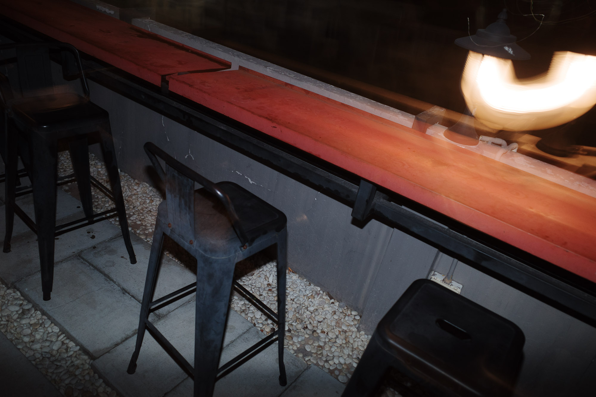 A dimly lit outdoor patio features industrial-style high stools arranged beside a long, narrow red countertop, with ambient overhead lighting casting warm glows onto the minimalist decor.