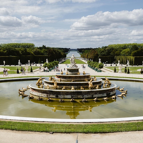 Una gran fuente ornamentada con estatuas doradas en un jardín formal, rodeada de senderos y personas, bajo un cielo nublado.