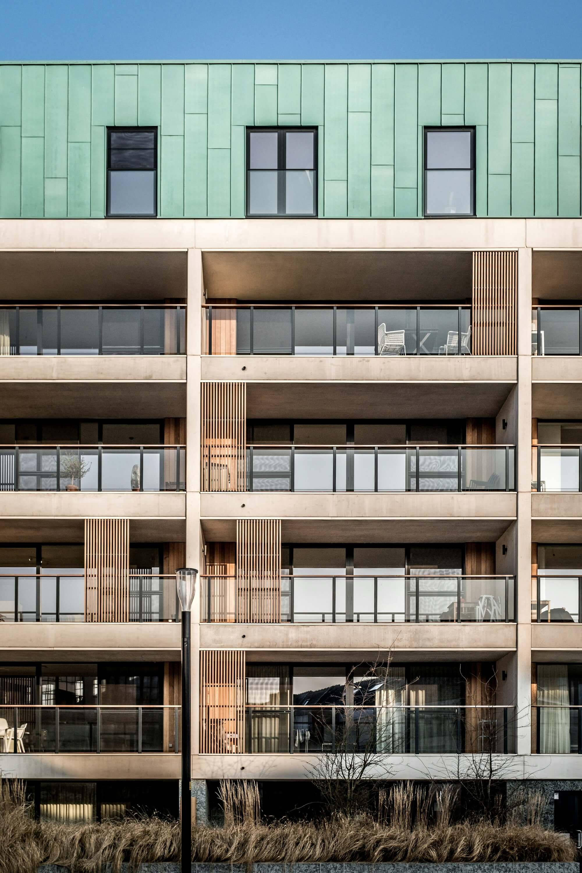 Modern apartment building with a green metal roof, large windows, and glass balconies. The structure features clean lines, wood accents, and a serene ambiance.