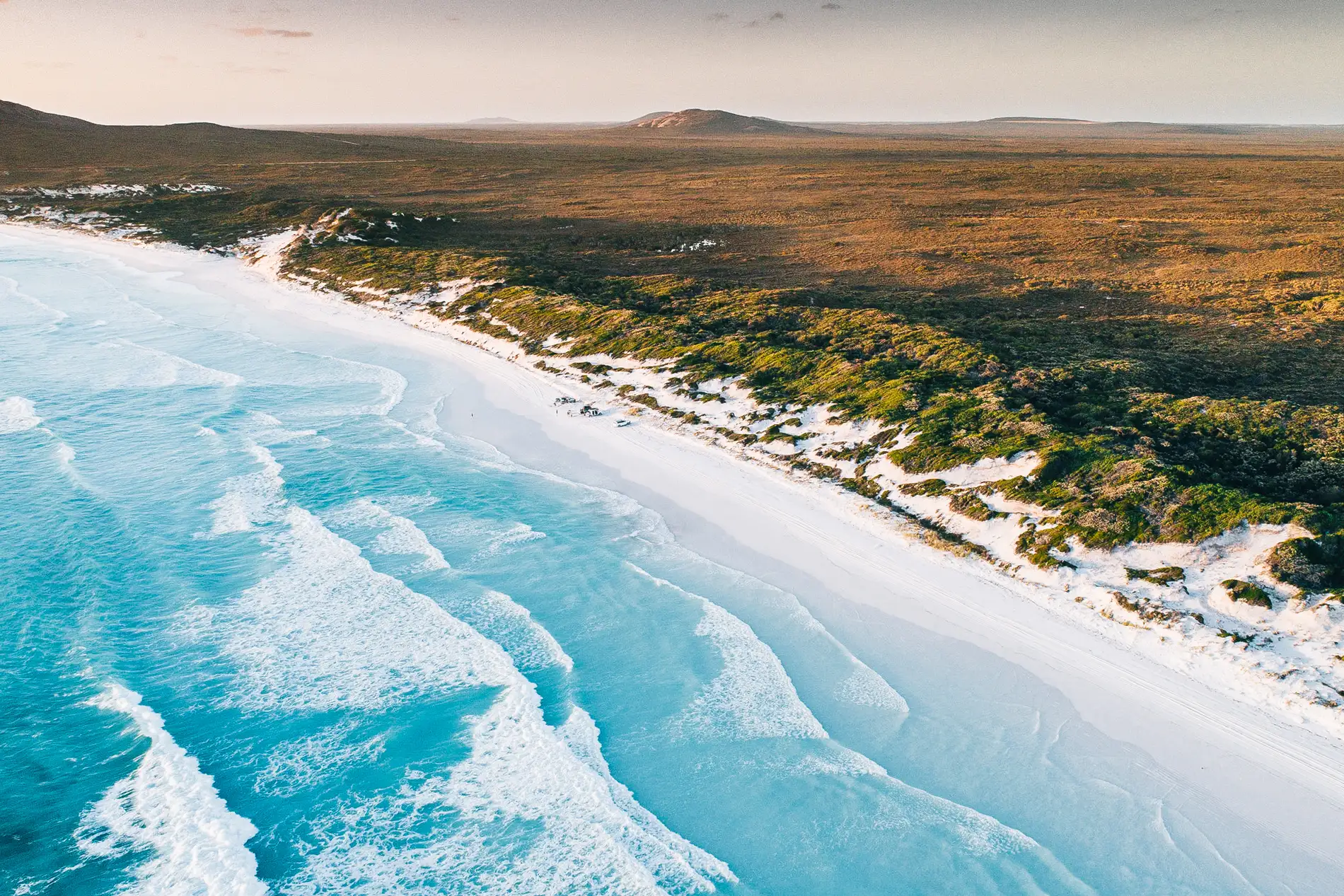 Aerial of a long white beach with lines of surf and low coastal scrub inland