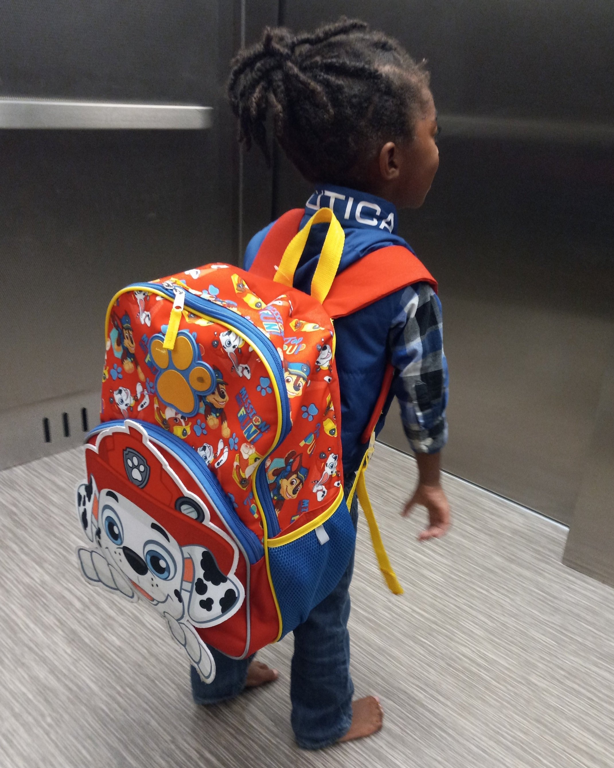 Alt tag: Young child standing barefoot in an elevator wearing a colorful character-themed backpack, facing the elevator doors.