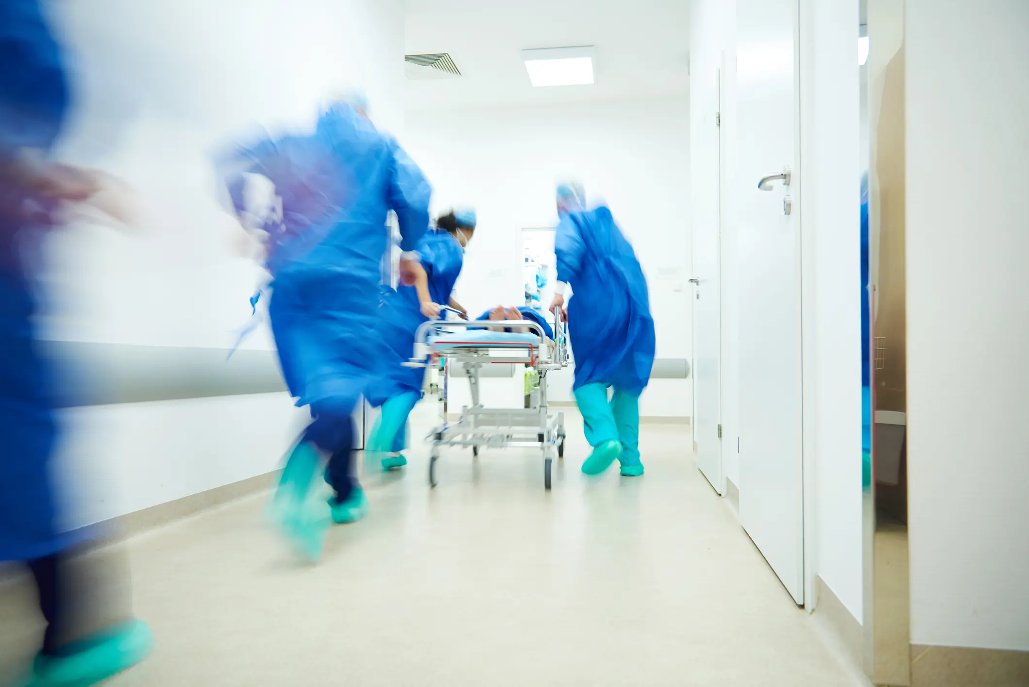 Blurred motion of healthcare staff in scrubs rushing a patient on a stretcher down a hospital corridor, illustrating downstream reactive work in health
