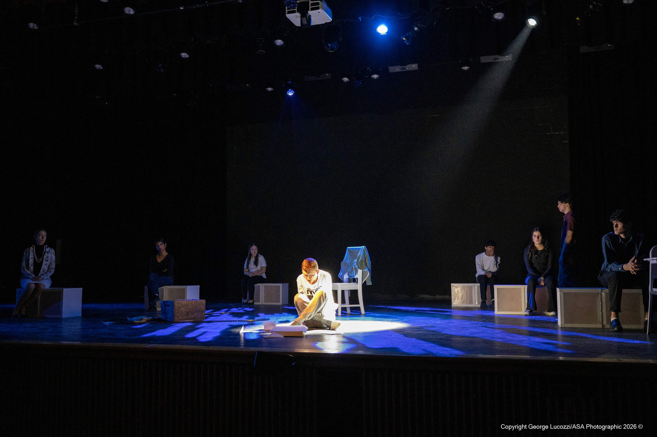 Christopher sits downstage center in a diagonal shaft of cool white light, while a deep indigo Cloud 15 gobo (Rosco 79065) washes the stage floor, creating a moody, atmospheric texture beneath him.