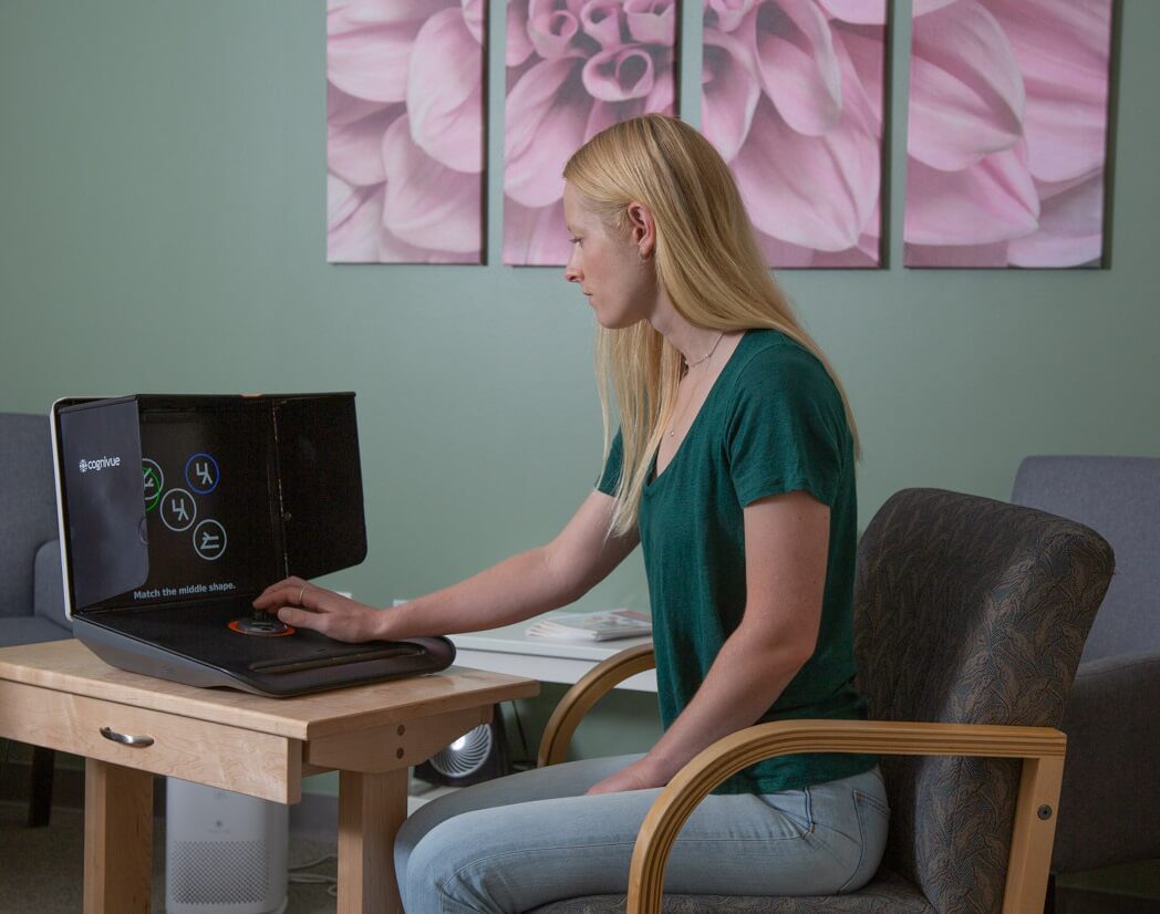 Woman with long blonde hair sitting at a desk, using a computer in a room with green walls and artwork.