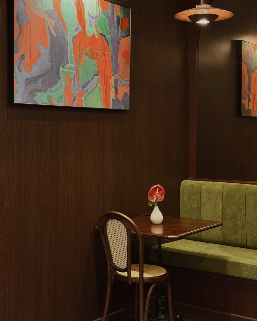 A cozy café setting featuring a wooden table and a chair with a woven backrest, beside a green upholstered bench. A small vase with an anthurium flower is placed on the table. The wall is adorned with colorful abstract artwork.