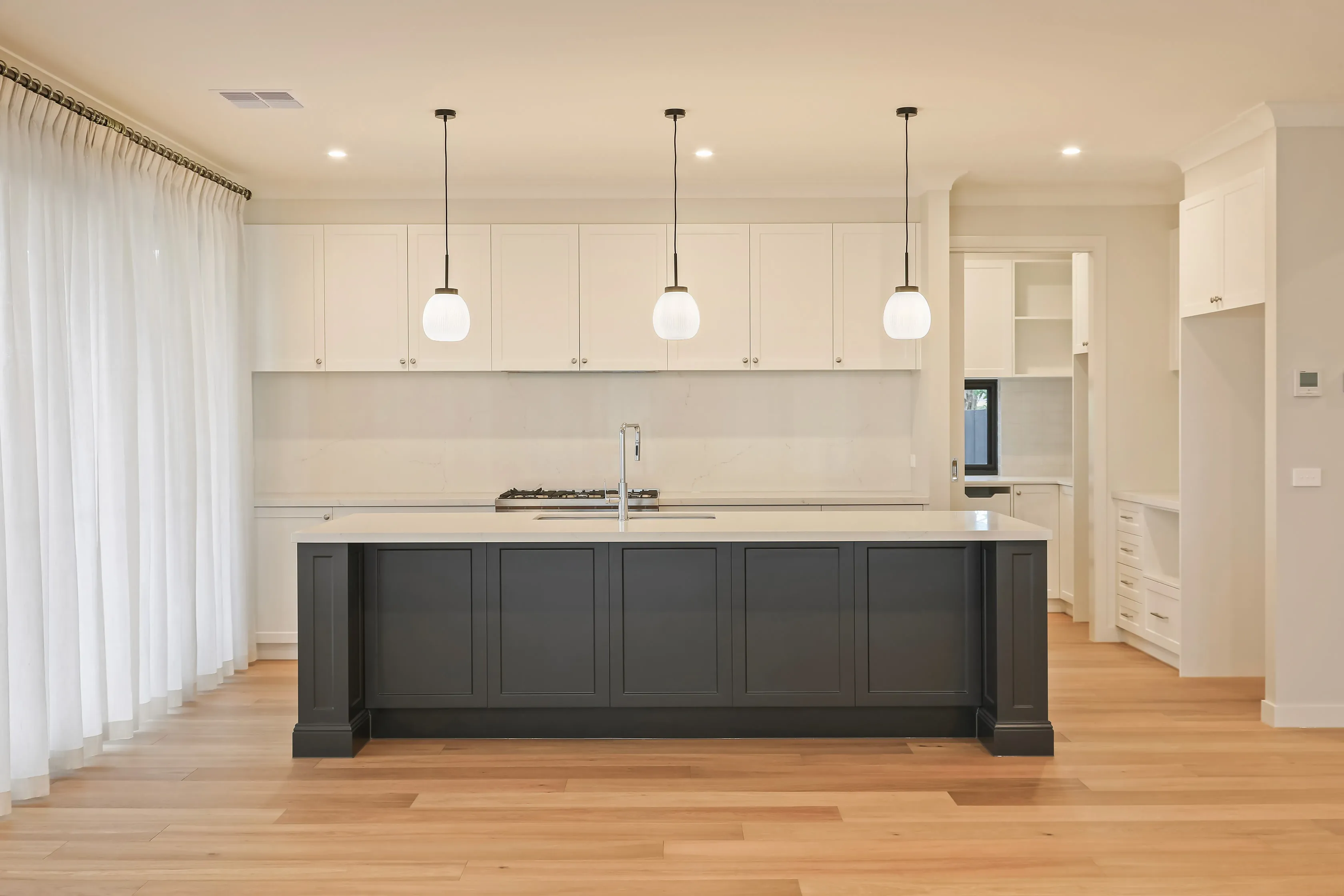 Kitchen with a long charcoal shaker island and white stone benchtop, three ribbed-glass pendant lights, white overhead cabinetry, gas cooktop, chrome tap and oak floors, with a butler's pantry beyond