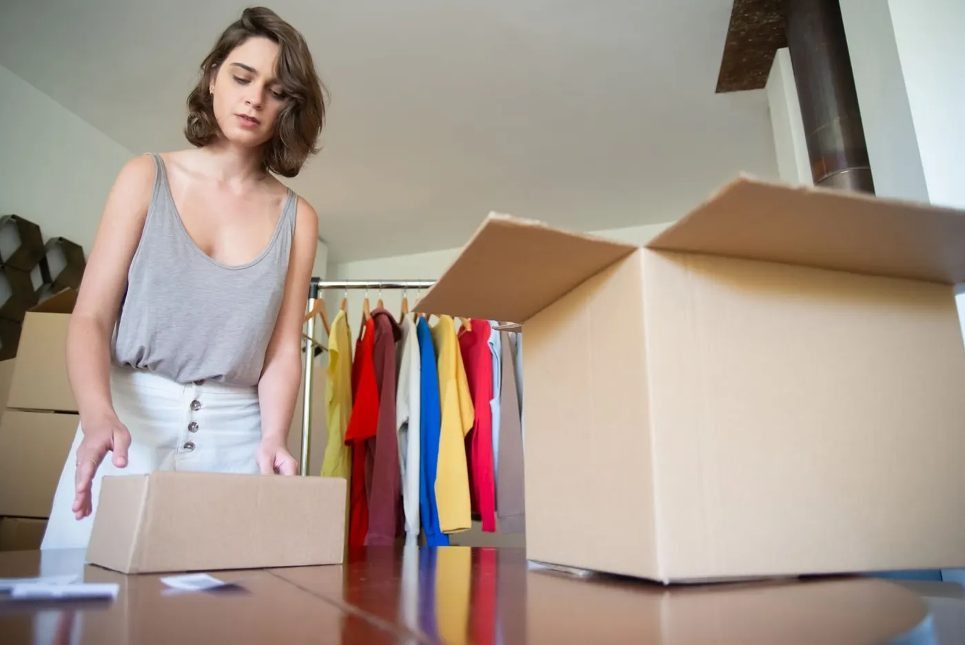 A woman packing an order into a cardboard box beside a rail of colourful shirts