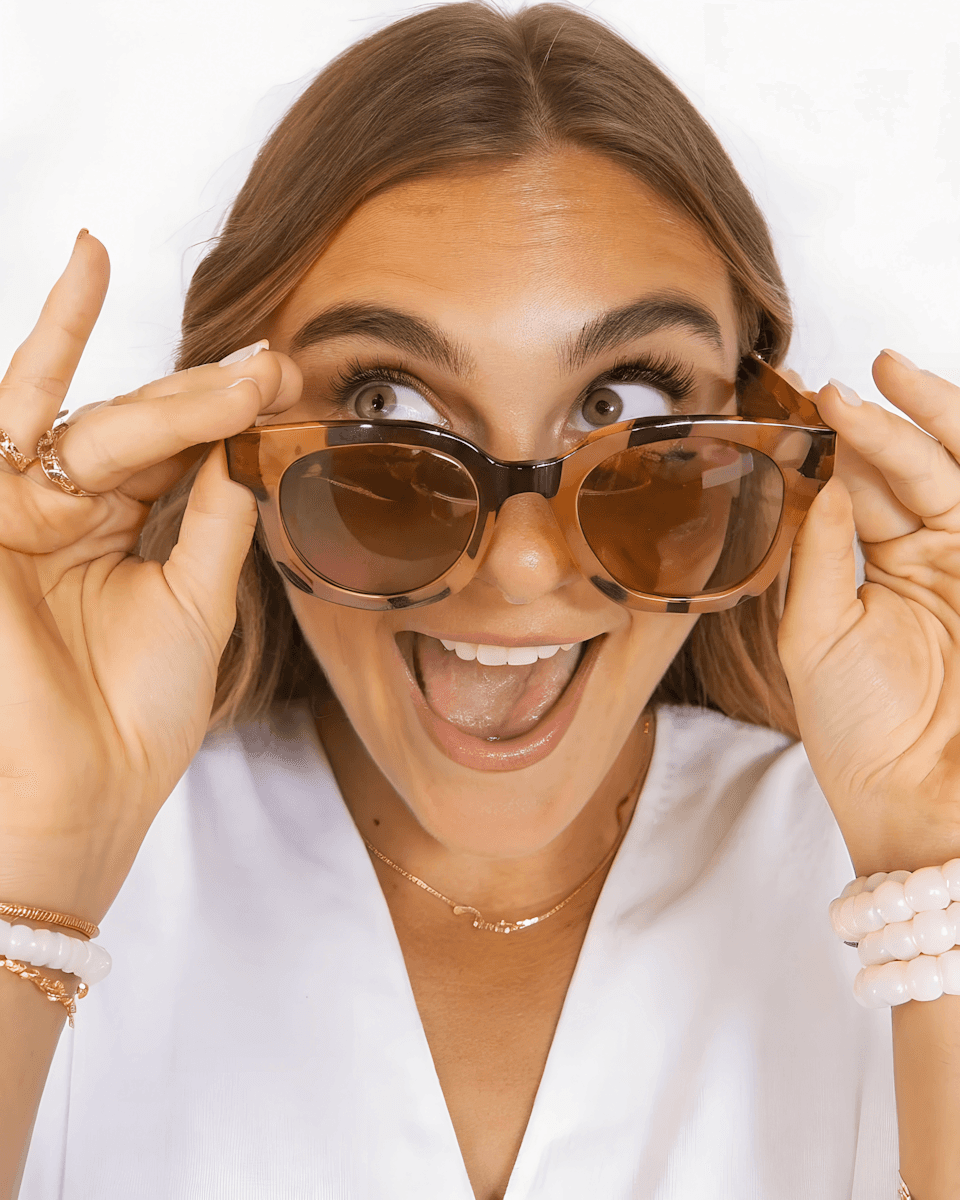 A young woman playfully adjusts her sunglasses, smiling and showcasing a cheerful expression against a light background.