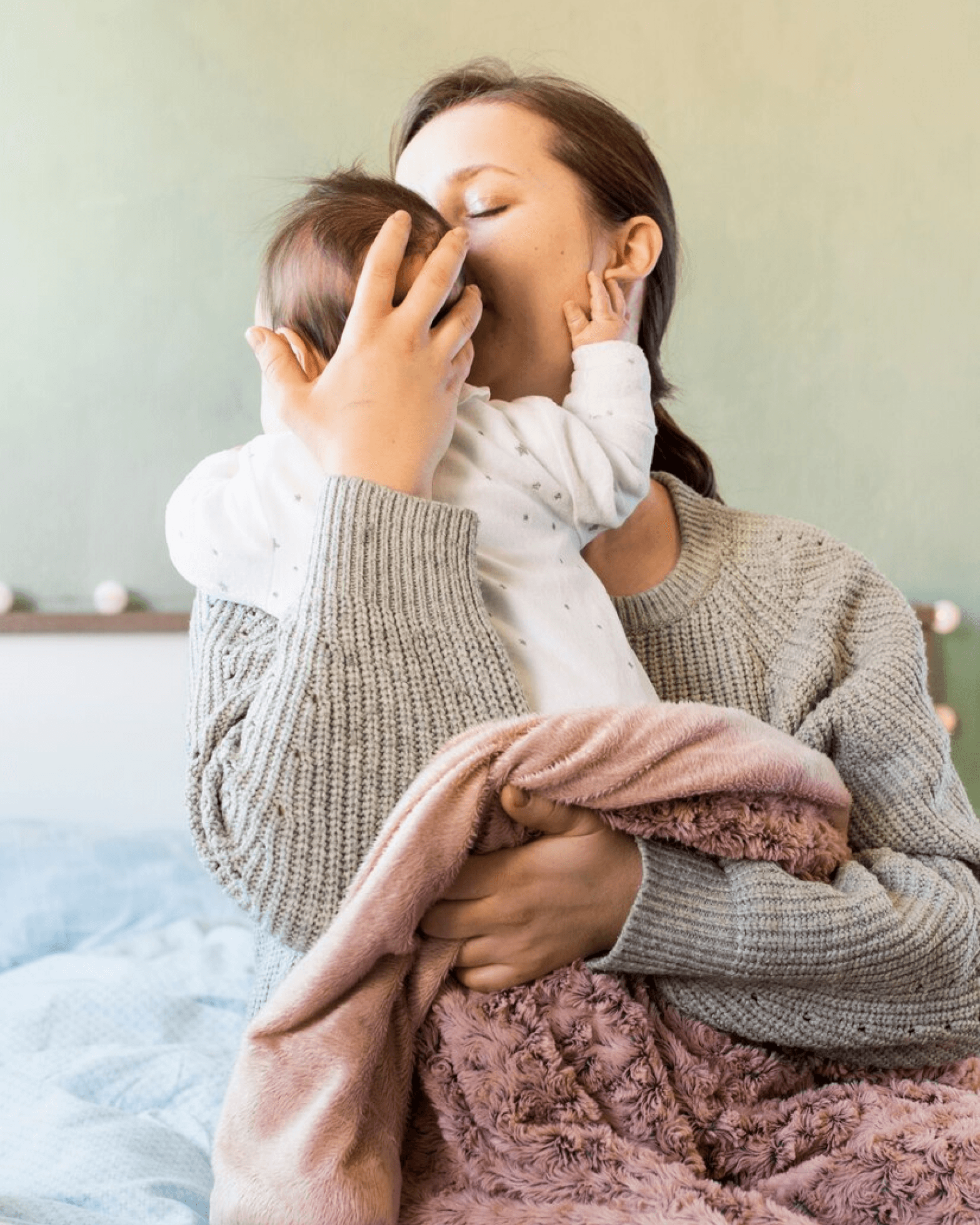 A serene mother in a cozy, gray sweater cradles her baby wrapped in a soft, pink blanket, exemplifying tender ayurvedic postpartum care in a peaceful bedroom setting.