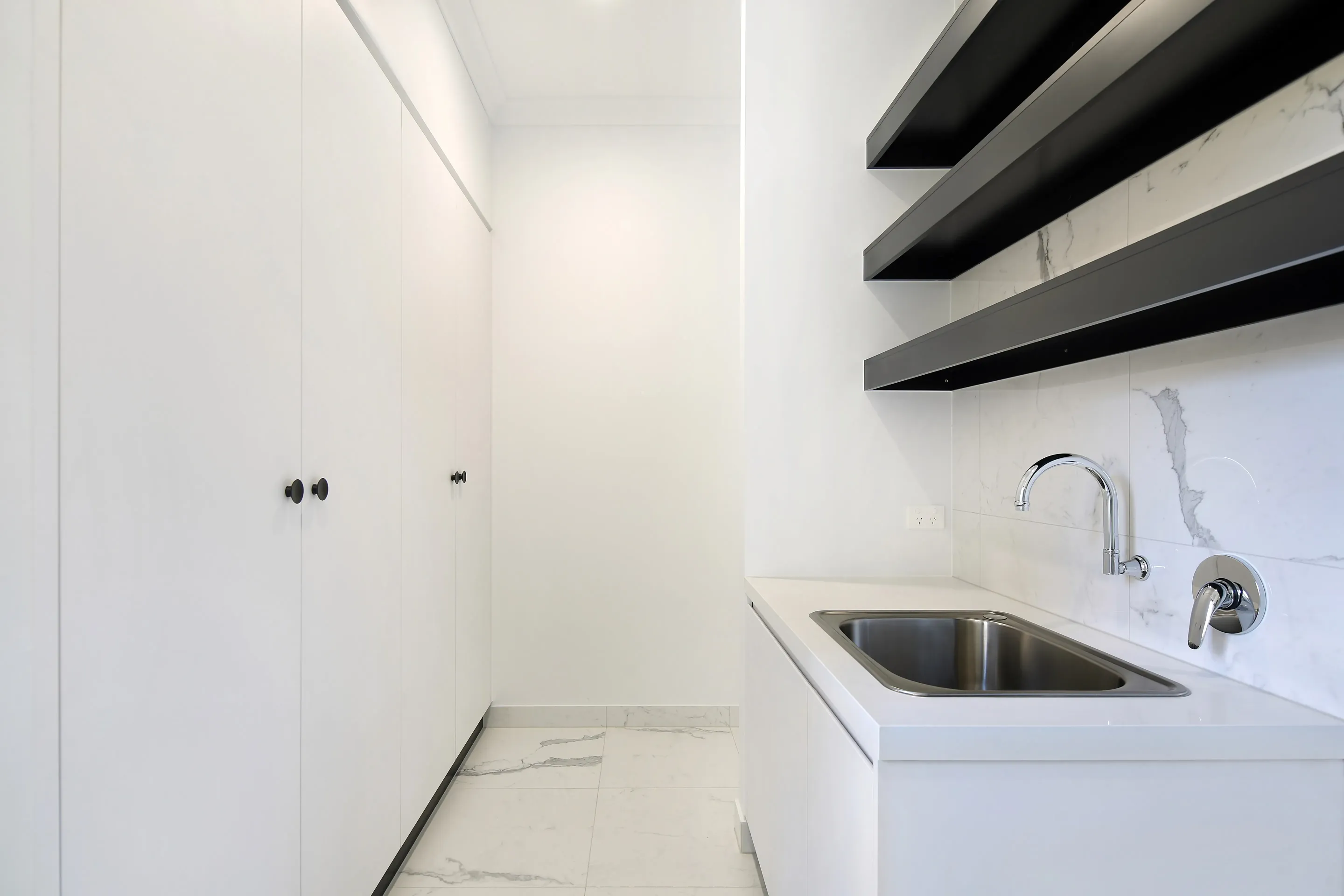 Laundry with white floor-to-ceiling cabinetry, black floating shelves, a marble-look splashback, white benchtop with stainless sink and chrome tap over marble-look floor tiles