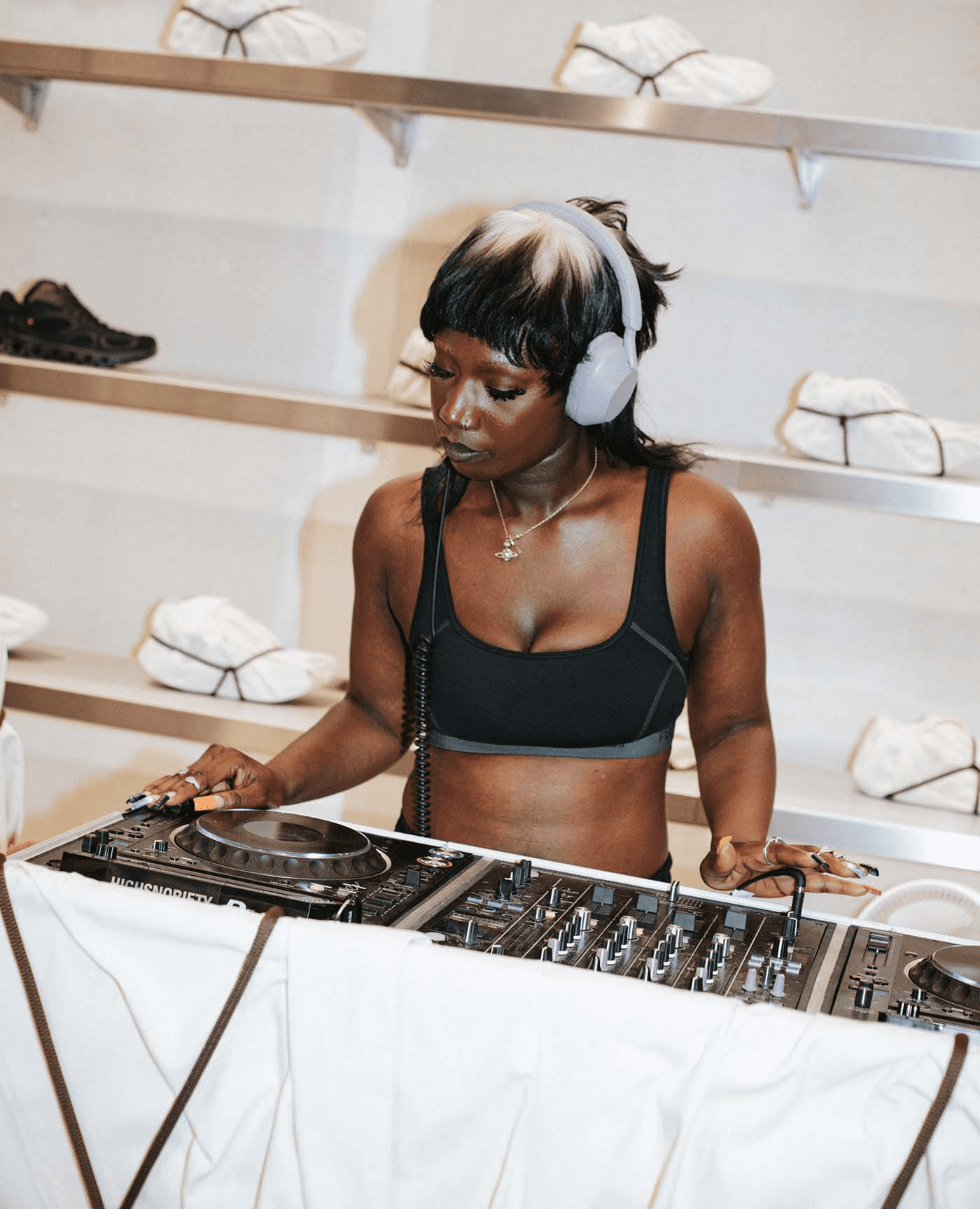A woman wearing headphones and a black sports bra is DJing at a mixing console, with shelves containing white items and a single black item in the background, showcasing her unique branding style.