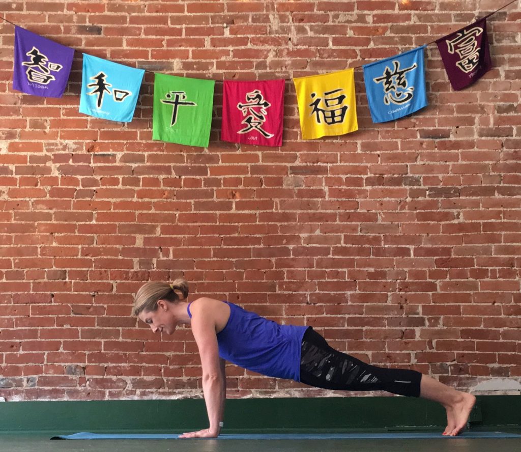 Heather Hausman practices a yoga plank pose on a blue mat against a brick wall adorned with colorful Tibetan prayer flags.