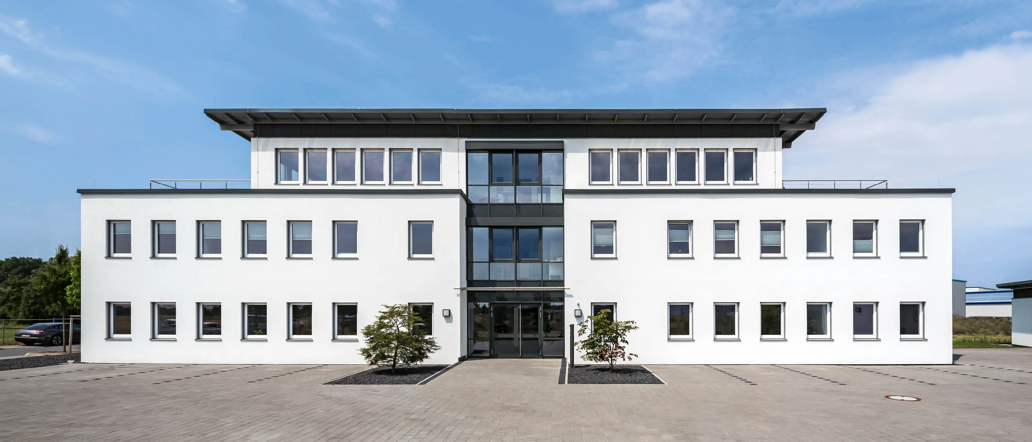 Modernes weißes Bürogebäude mit großen Fenstern und einem Vordach über dem Eingangsbereich, auf einem gepflasterten Vorplatz.