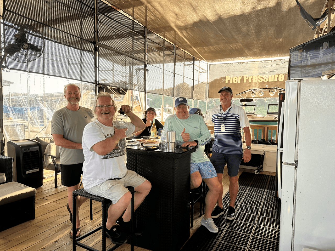 Four men are standing and sitting around a black bar on a boat deck, smiling and enjoying drinks and snacks, with a background of a marina and a sign that says "Pier Pressure" under a canopy.