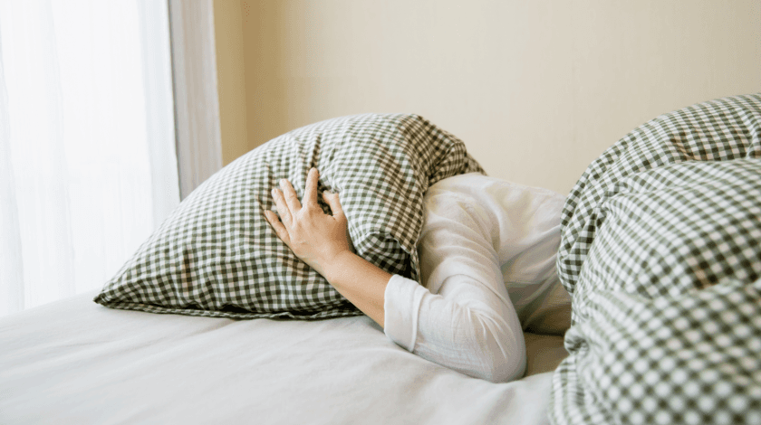 A person covers their head with a checkered pillow while lying in bed, creating a cozy and restful atmosphere in a softly lit room.