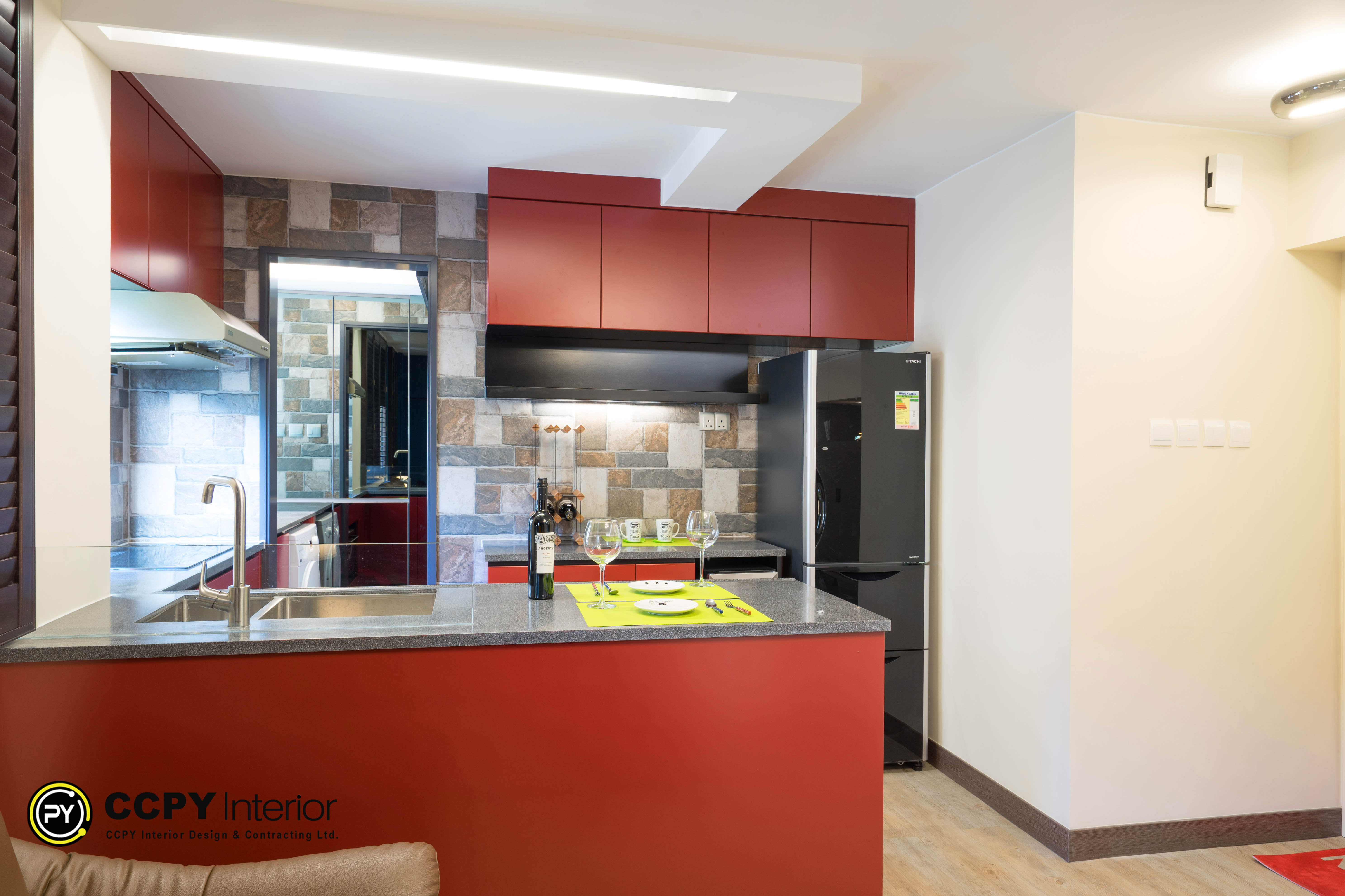 his is a red and black modular kitchen.The kitchen features a dark grey countertop and red cabinets.It is equipped with a black refrigerator and backsplash tiles in varied neutral tones.The space is styled with an island setup, including a bottle of wine and glasses on the counter.