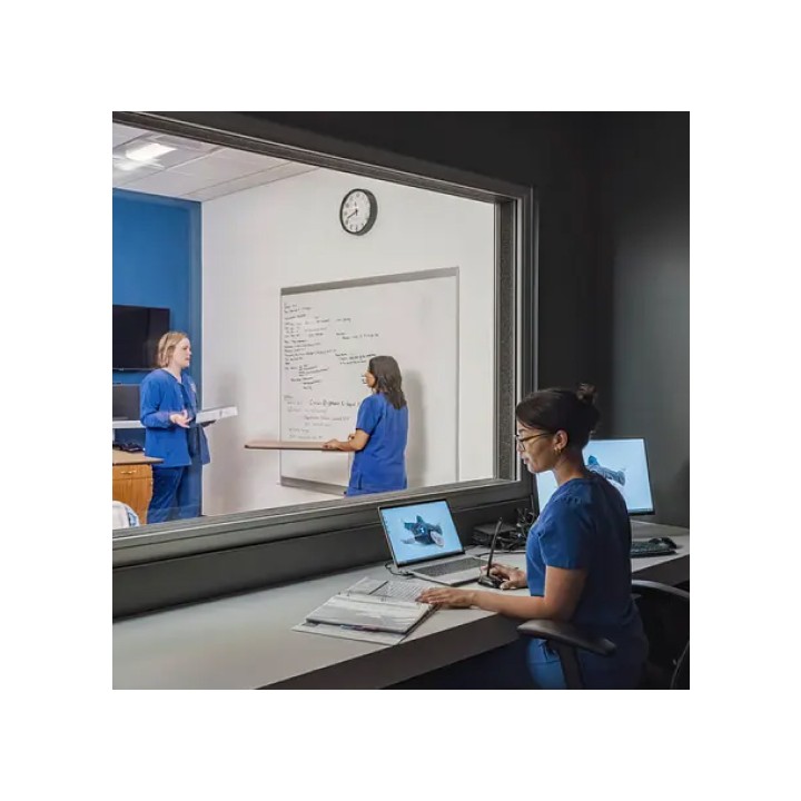 A nurse in a control room observes two medical professionals in blue scrubs discussing notes on a whiteboard. The setting is focused and professional.