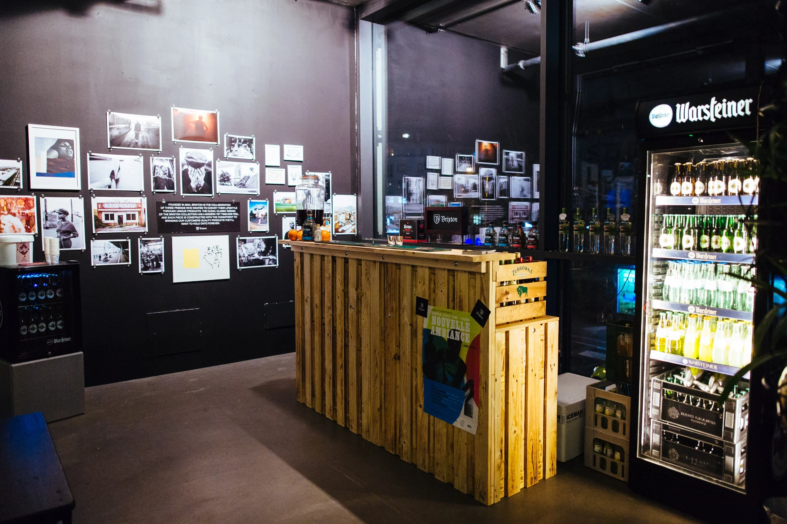 Pallet-wood bar at the BRIXTON pop-up with a drinks dispenser, a Warsteiner fridge full of bottles and a black wall covered in framed photography.
