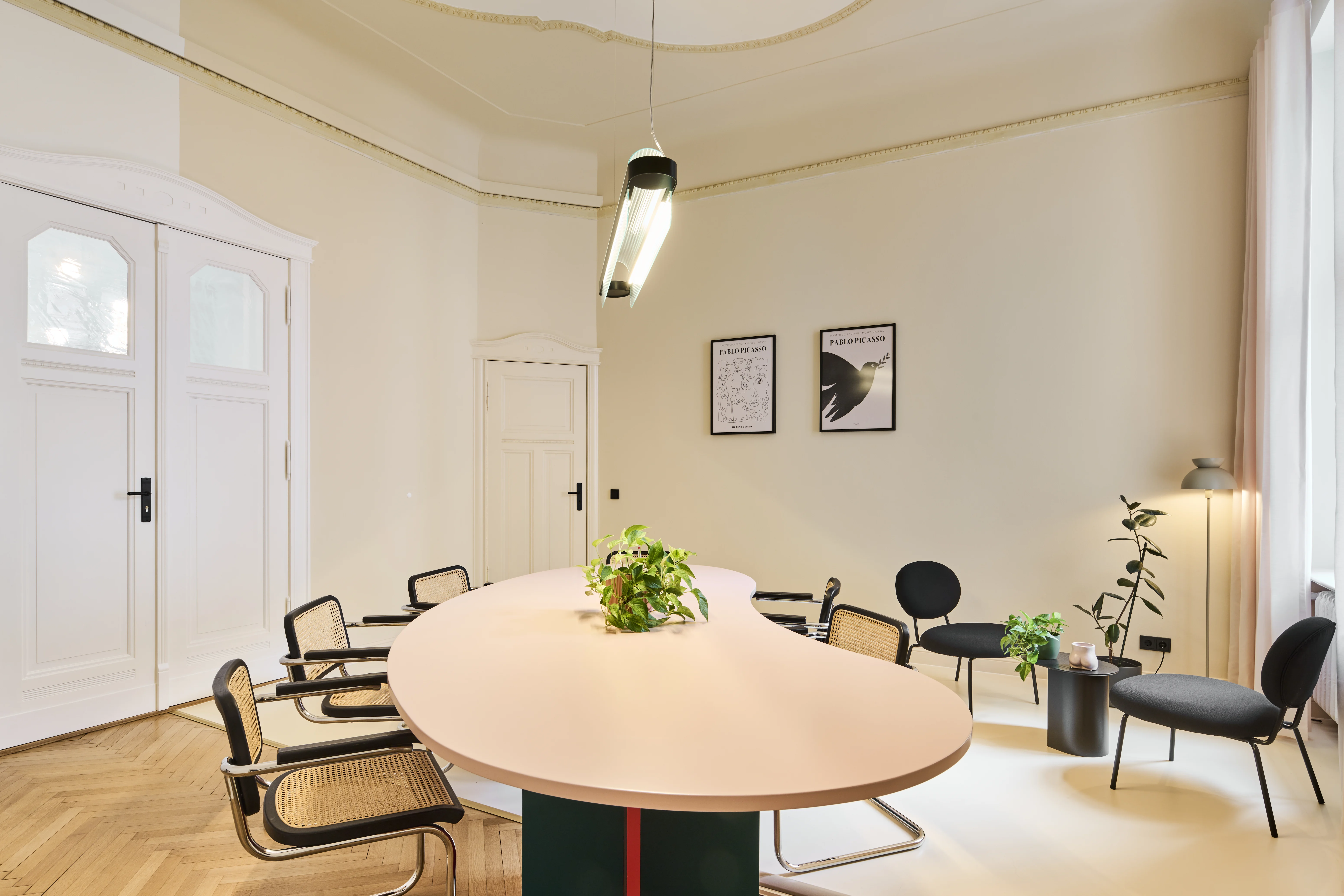 Conference room at INTERMATE BERLIN with a curved blush-pink table on a green base, cane-back cantilever chairs, black chairs and side table, posters and a tubular pendant light.