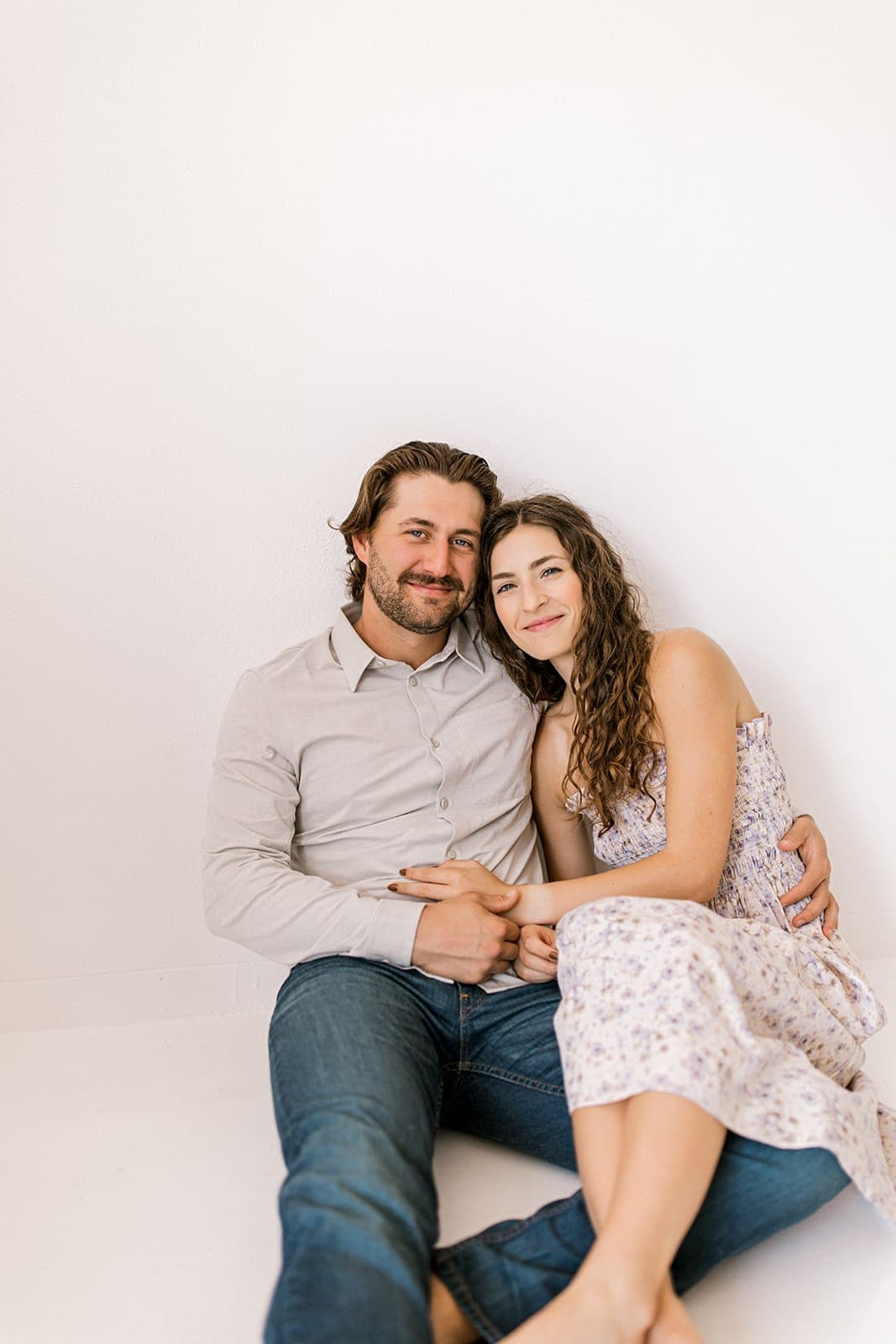 A couple sits on the floor, smiling at the camera during their natural light engagement session at Revelator Studio, Shreveport.