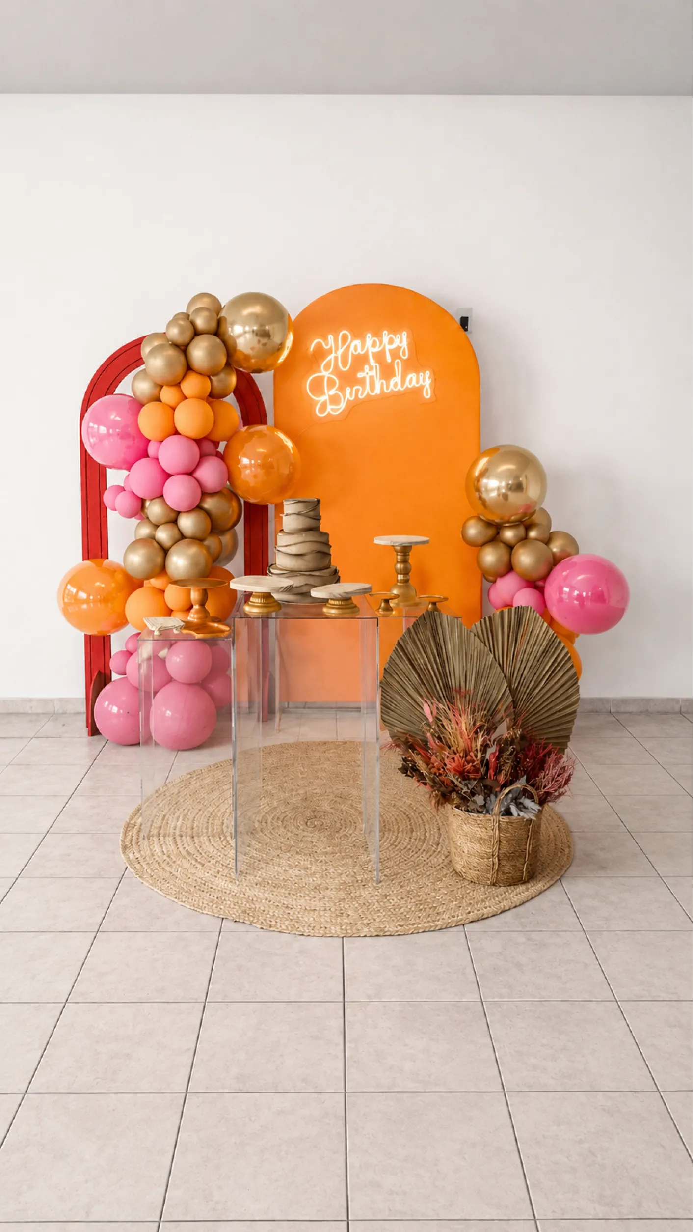Decoração tropical com painel laranja, balões rosa, laranja e dourado, letreiro Happy Birthday, mesa transparente e arranjos naturais.