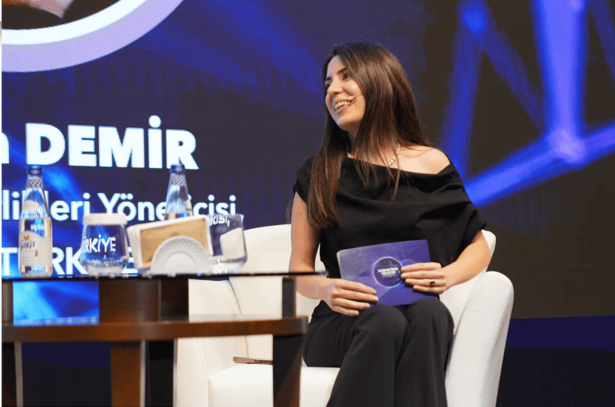 Öykü’nün sahnede bir koltukta oturduğu, elinde sunum metninin bulunduğu kartları tuttuğu ve mikrofonla konuştuğu bir sahne.