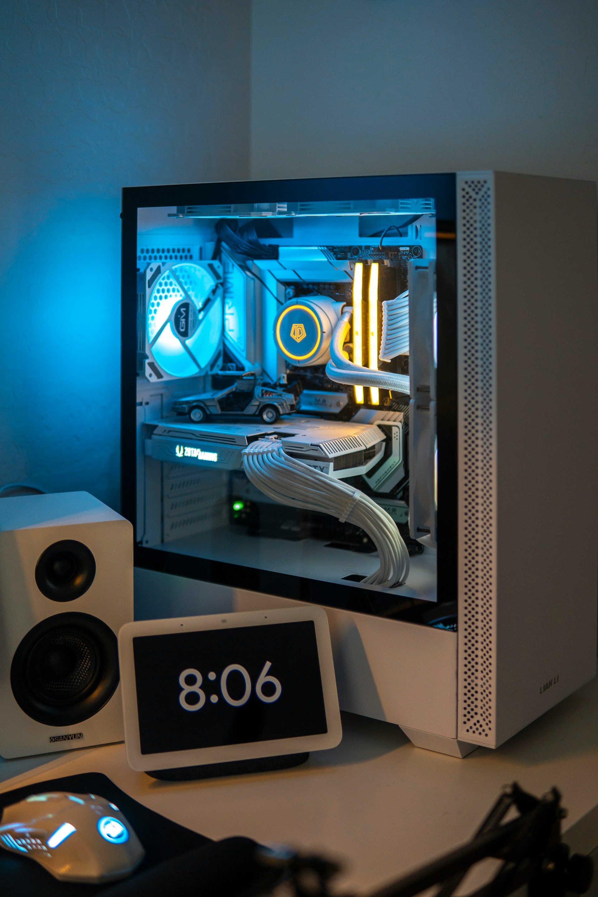 A white custom-built desktop computer case with blue and white internal lighting, sitting next to a white smart display showing "8:06" and a speaker.