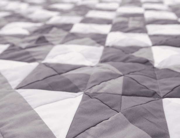 A close-up view of a quilted blanket or bedspread. The quilt features a geometric pattern of interlocking triangles in shades of gray and white. The fabric has a slightly textured appearance with visible stitching. The image focuses on the quilt’s pattern and texture, without any additional elements or context.