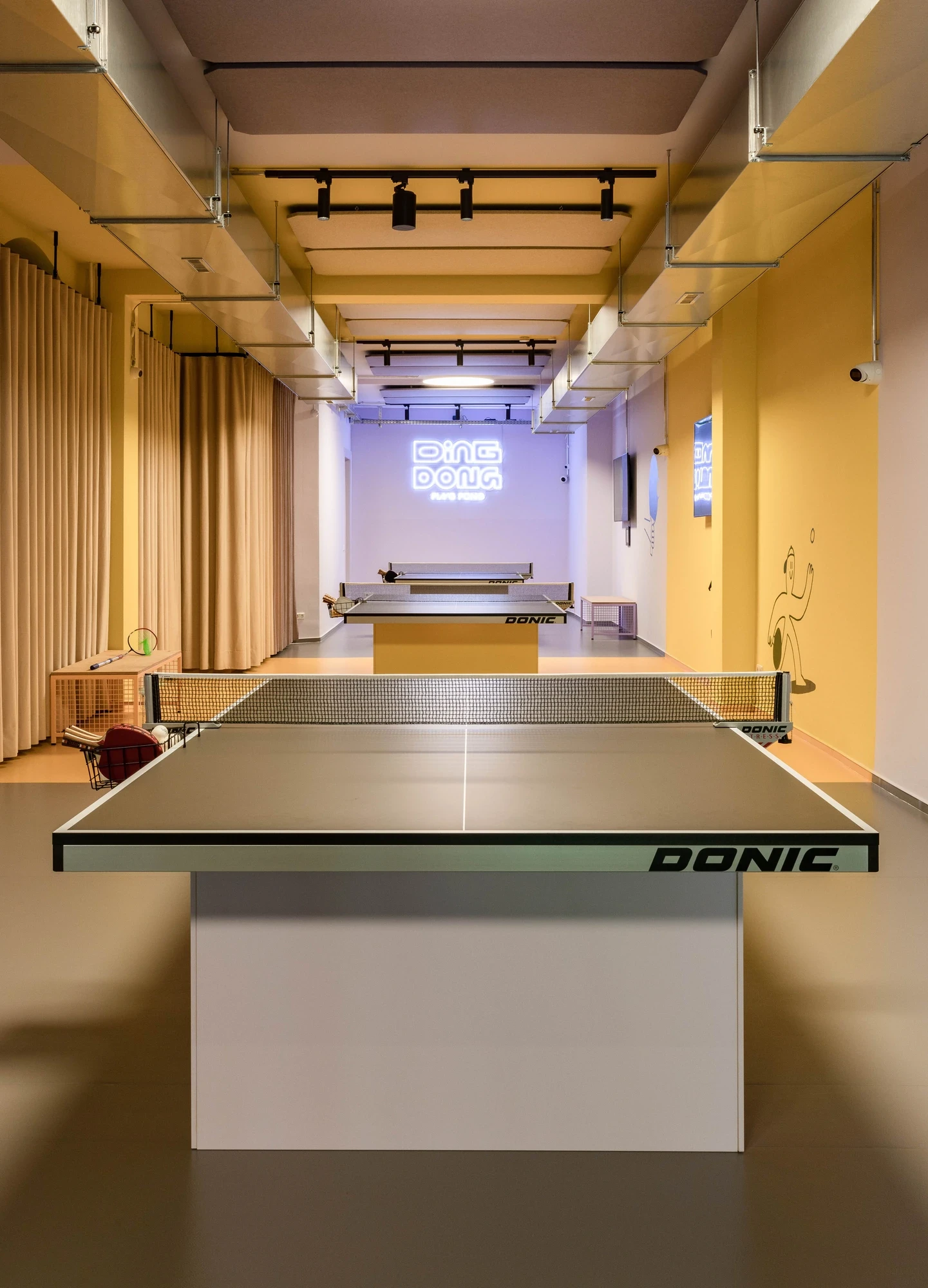 DingDong ping pong room with two Donic table tennis tables in a corridor of warm yellow walls and a glowing „DingDong Ping Pong" sign on the back wall.