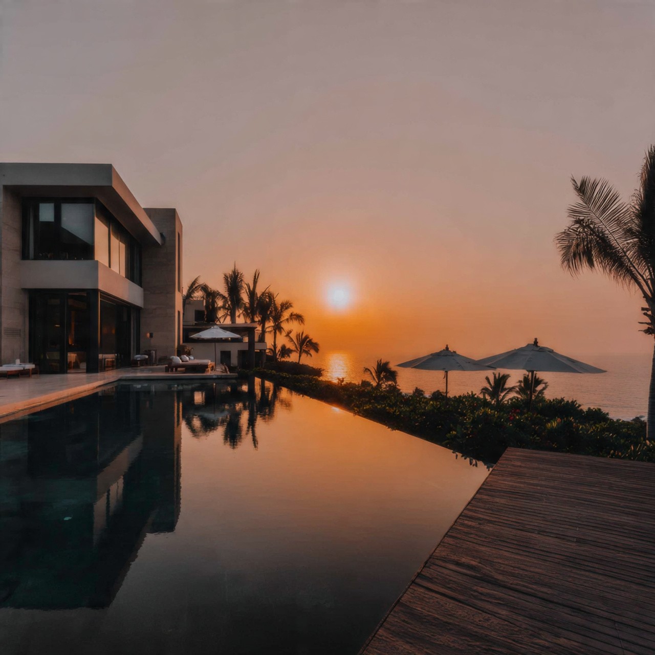 Modern villa at sunset overlooking an infinity pool reflecting the orange sky. Palm trees and umbrellas create a serene, tropical atmosphere.