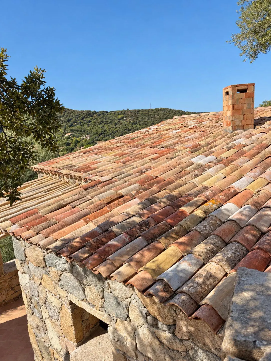 Toiture en tuiles canal de récupération aux nuances variées et souche de cheminée en terre cuite sur murs en pierre