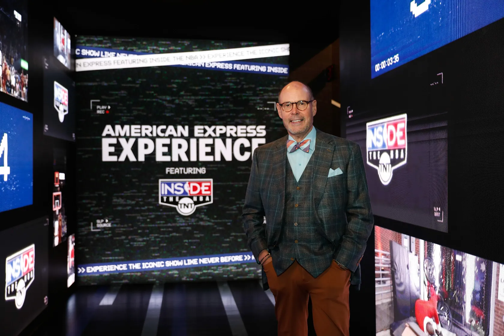 Ernie Johnson in a plaid jacket, bow tie, and glasses stands smiling in front of a large screen displaying American Express Experience featuring Inside the NBA in a modern, well-lit event space.