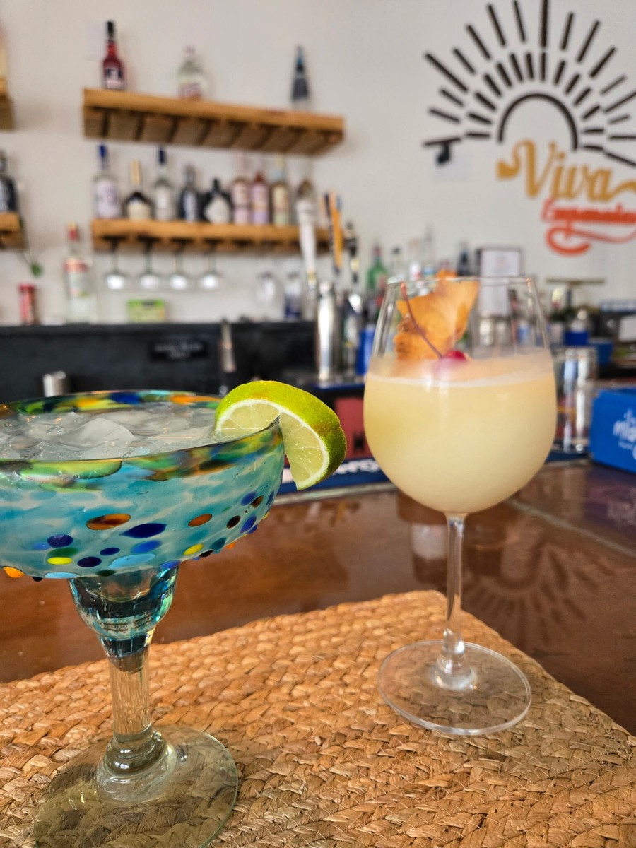 Two colorful cocktails on the bar, with the Viva sun logo and shelves of bottles behind