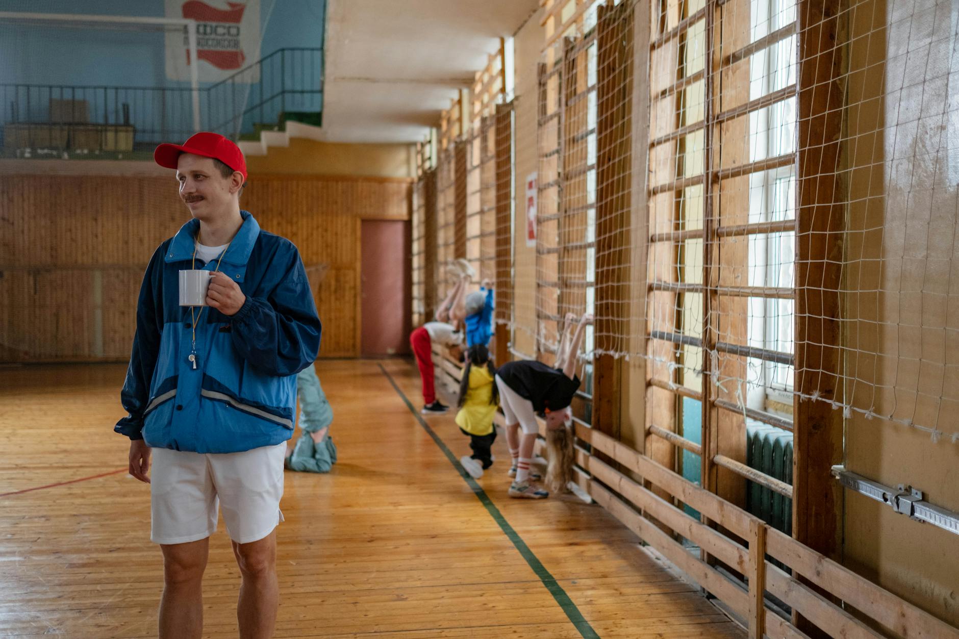 A physical education teacher stands in a bright gym holding a clipboard and whistle while greeting students.