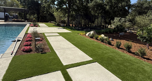 Artificial grass poolside lawn in Loomis with concrete step-stones, flowering border plantings, and boulder accents under oaks.