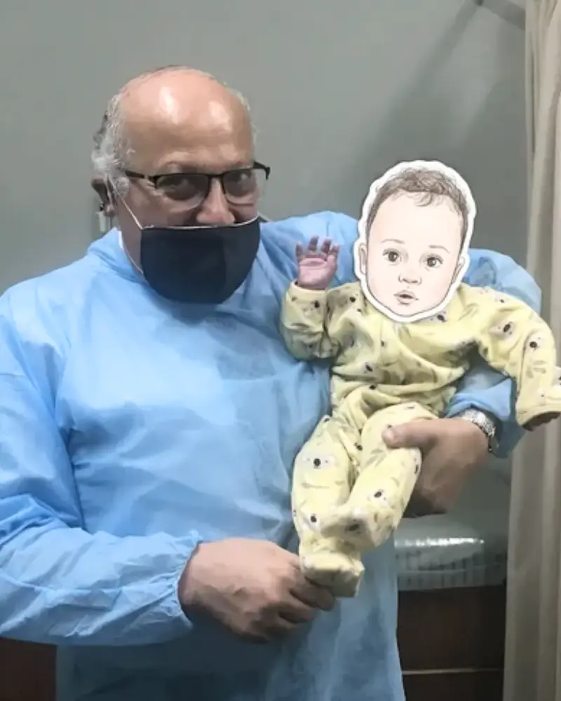 Dr. Ashraf Saddik holding a baby in a yellow patterned onesie raising his hand inside a Coburg clinic room.