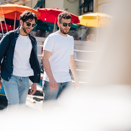 Dva muži nosí sluneční brýle a neformální oblečení, jak jdou venku, zatímco v pozadí jsou jasně zbarvené deštníky.