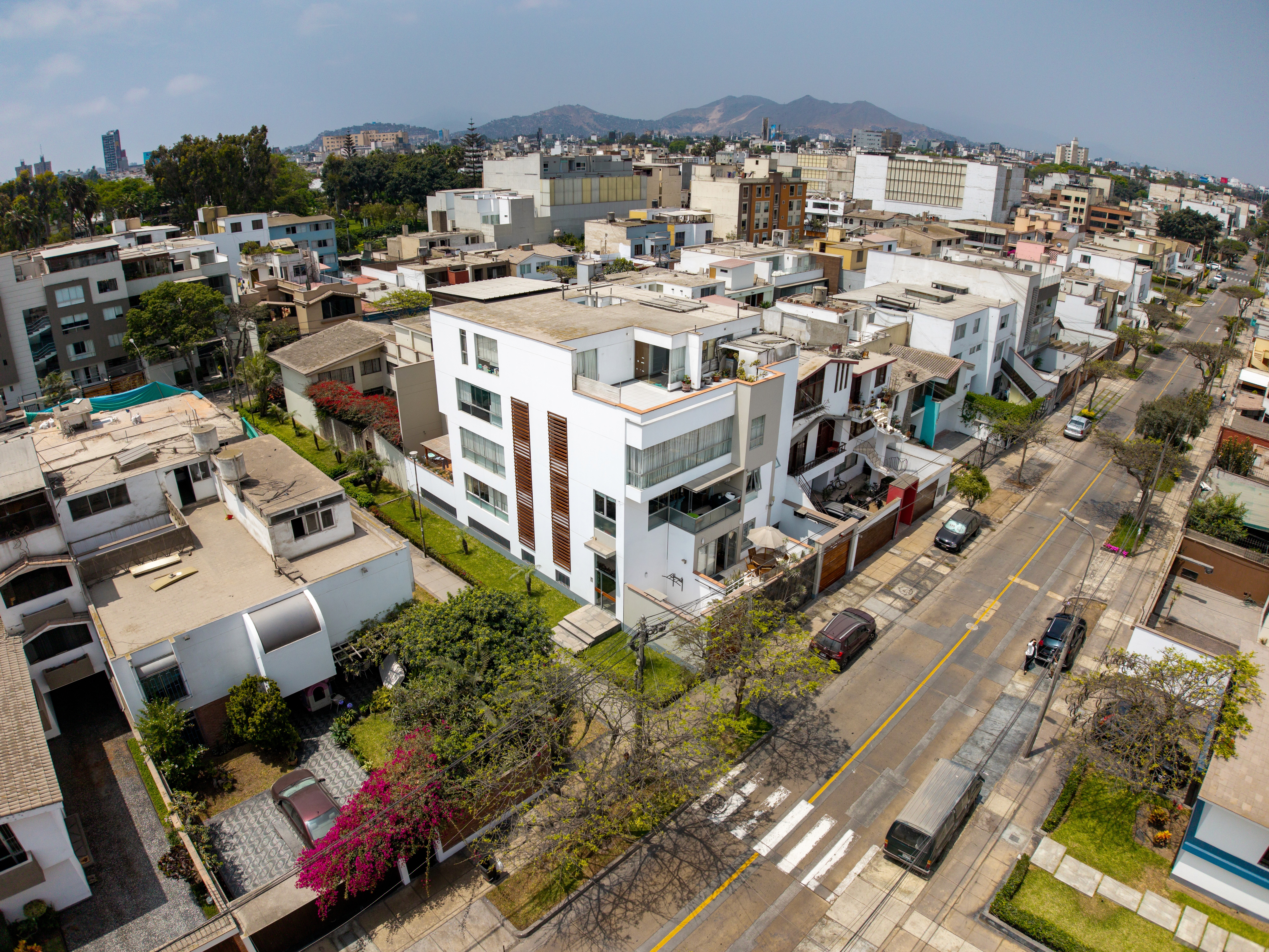 Aerial view of a geometric plaza with crisscrossing red paths and grass, surrounded by white buildings and a tree-lined street with cars. Urban, organized.