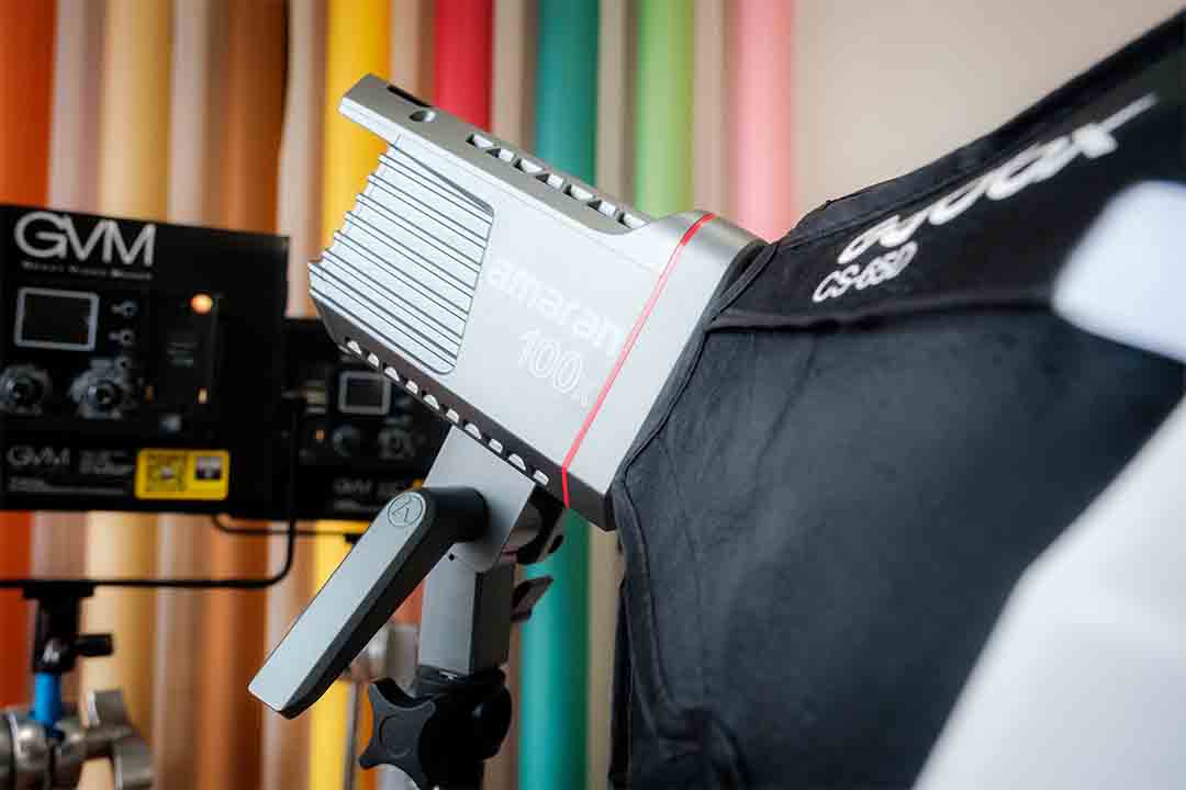 Close-up of a professional Amaran 100x studio light, with additional lighting equipment in the background against colorful backdrop rolls in a photo studio.