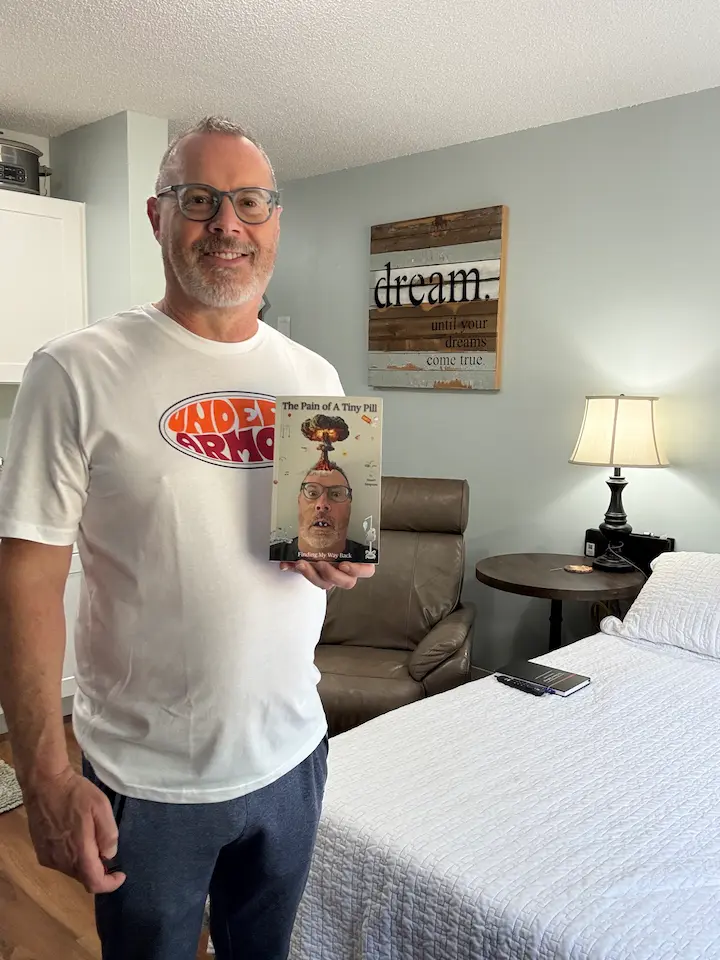 Stu in his living room holding his old book The Pain of A Tiny Pill.
