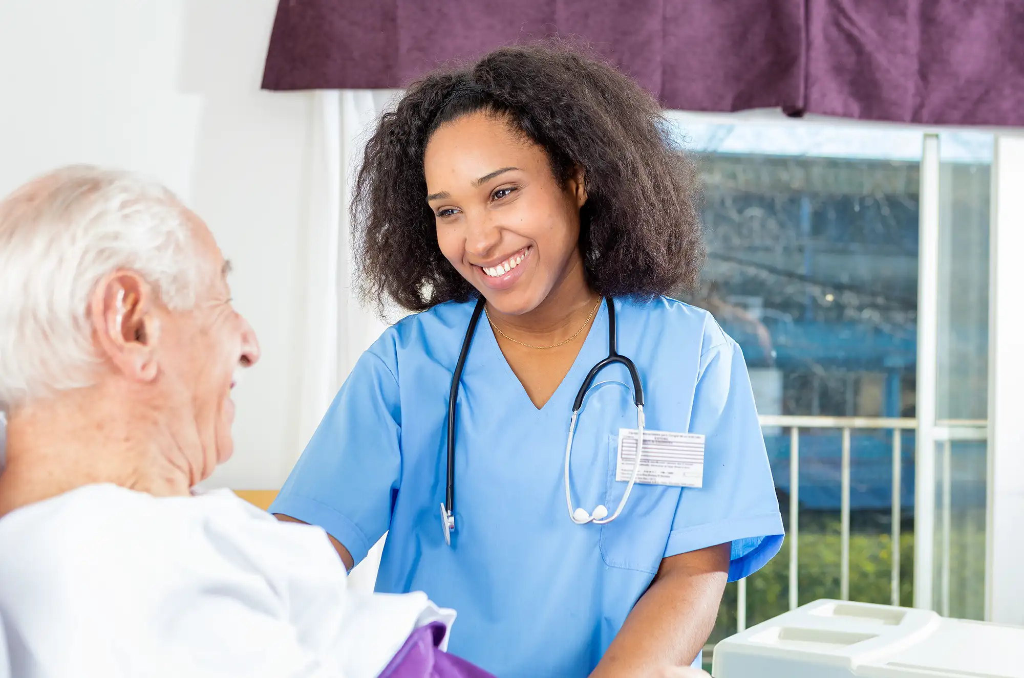 Smiling nurse interacting with an elderly patient in a bright room.