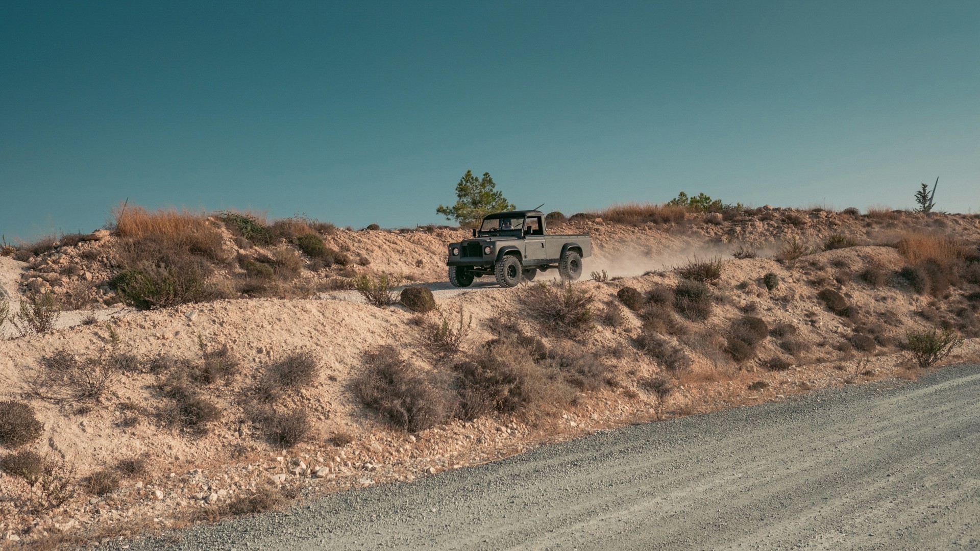 Dark grey Land Rover Series pickup driving along a dusty ridge track through arid Cyprus scrubland, kicking up dust, under a clear blue sky.