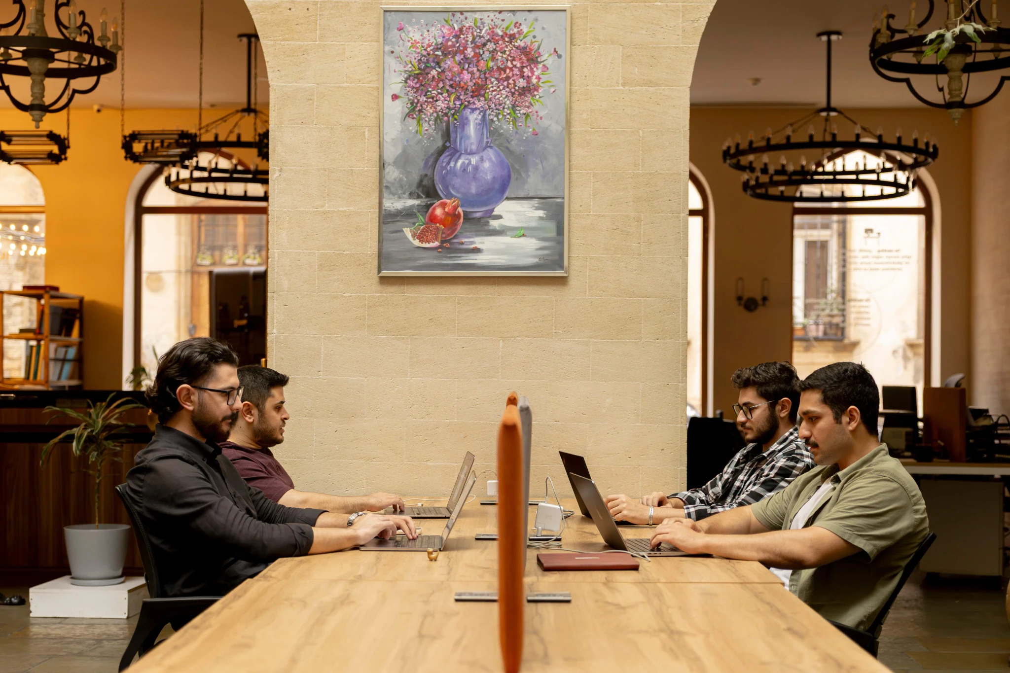 Open office scene: four colleagues typing on laptops across a long table, arch windows and wall art behind them.