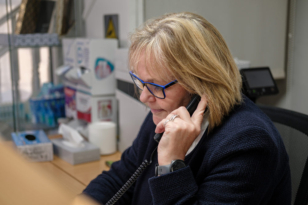 A woman with blonde hair and blue glasses is sitting at a desk speaking into a black corded phone, with office supplies and a stack of tissues visible in the background.