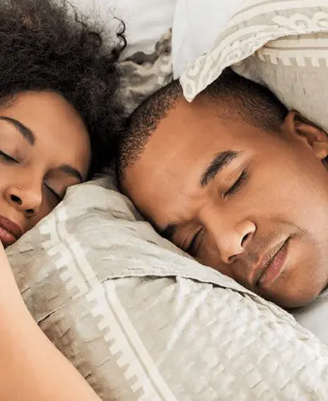 A couple is lying in bed, peacefully resting under a cozy blanket, smiling gently at each other.