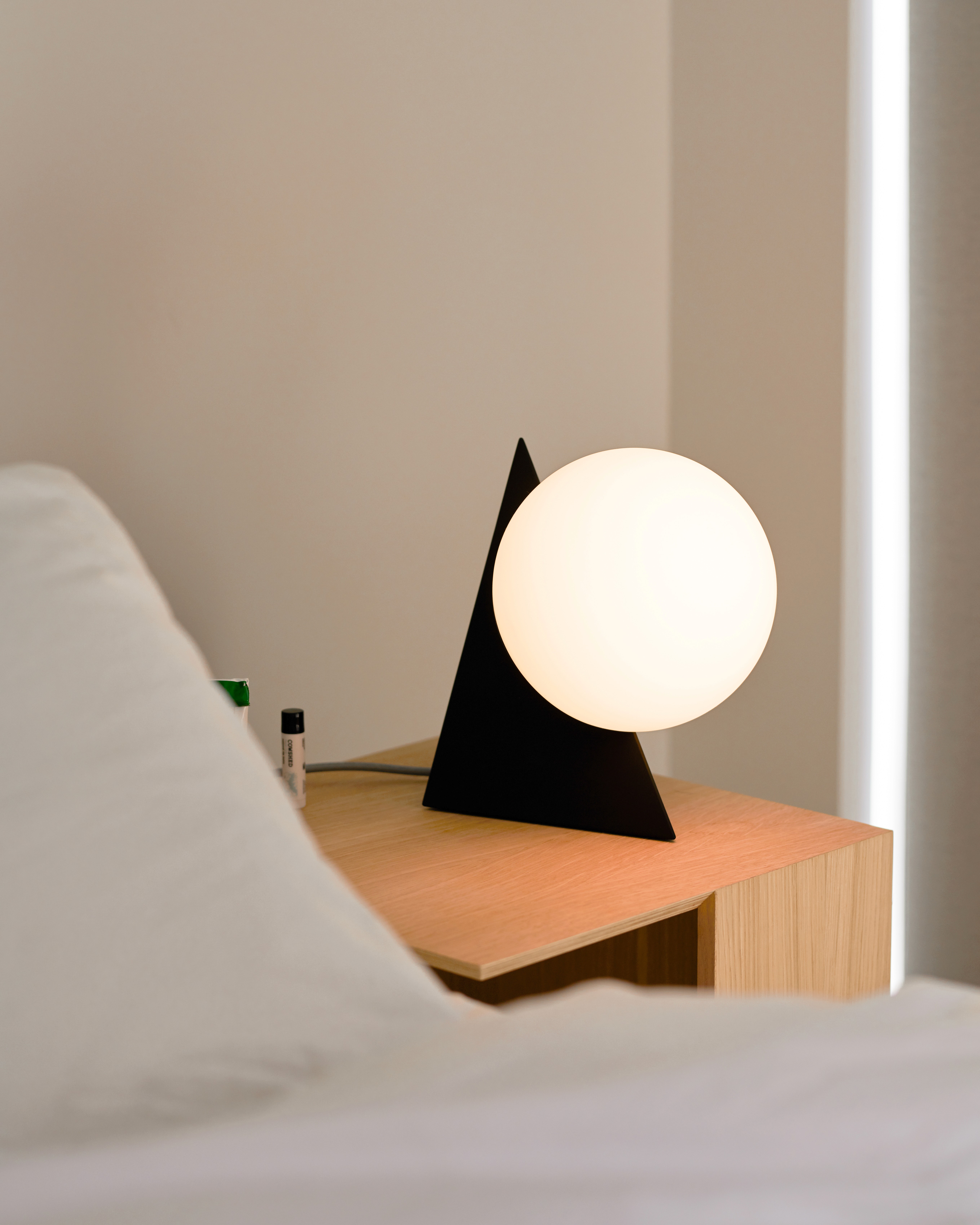 Black triangle table lamp holding a glowing white globe, on a wood nightstand beside a bed