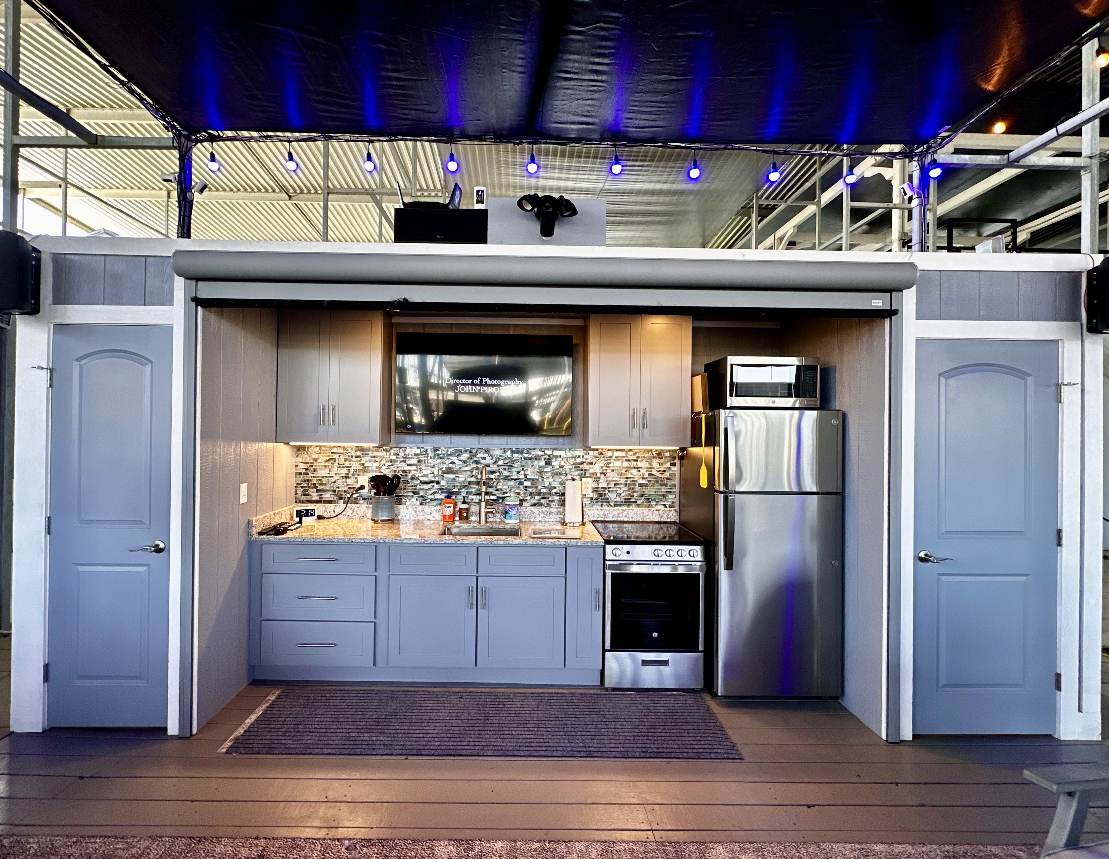 A modern outdoor kitchen features sleek gray cabinetry, a high-tech entertainment system, stainless steel appliances, and a stylish black ceiling with blue accent lighting.