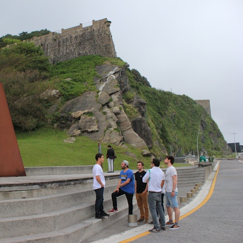 A group of five men casually conversing on steps near a hillside fortress with a rocky terrain beneath a cloudy sky.